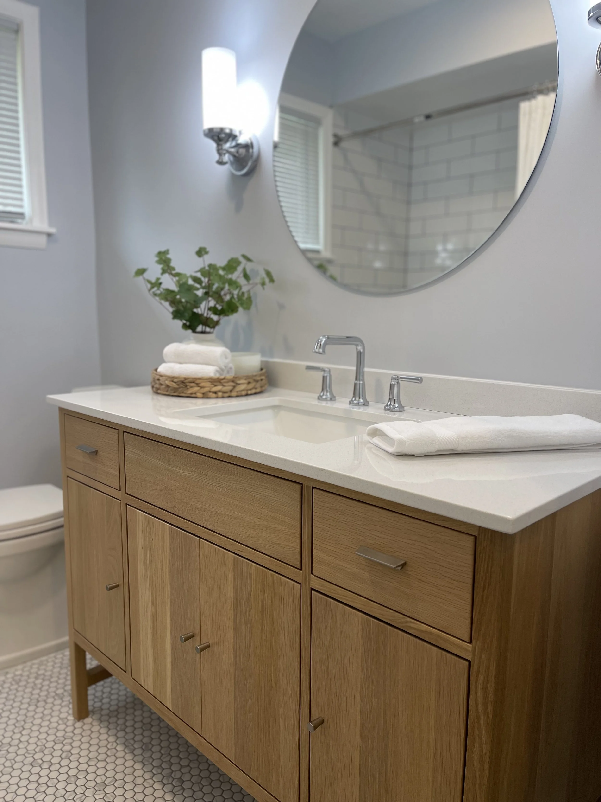Bathroom vanity with wooden cabinet, white countertop, and silver faucet, decorated with a basket holding towels and a potted plant. A large mirror is above the sink, and wall-mounted lights are flanking the mirror. Part of the toilet and window with
