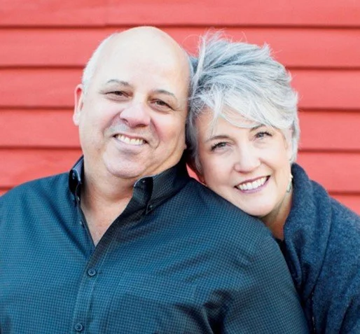 A smiling older couple, a man and a woman, standing close together against a red wooden wall. The man has a bald head and is wearing a dark blue shirt. The woman has short gray hair and is wearing a dark sweater.