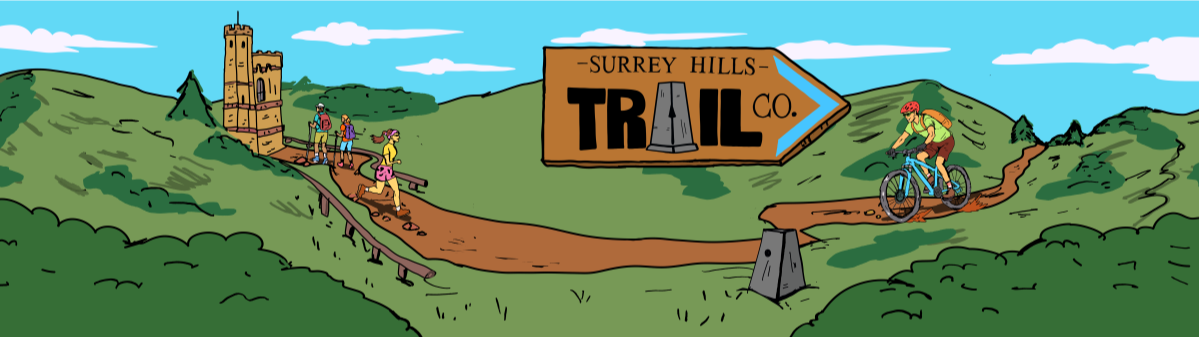 A scenic trail in Surrey Hills with hikers walking and a cyclist riding a mountain bike along a dirt path, with a wooden trail sign pointing toward Surrey Hills Trail Co., and a castle-like structure on a hill in the background.