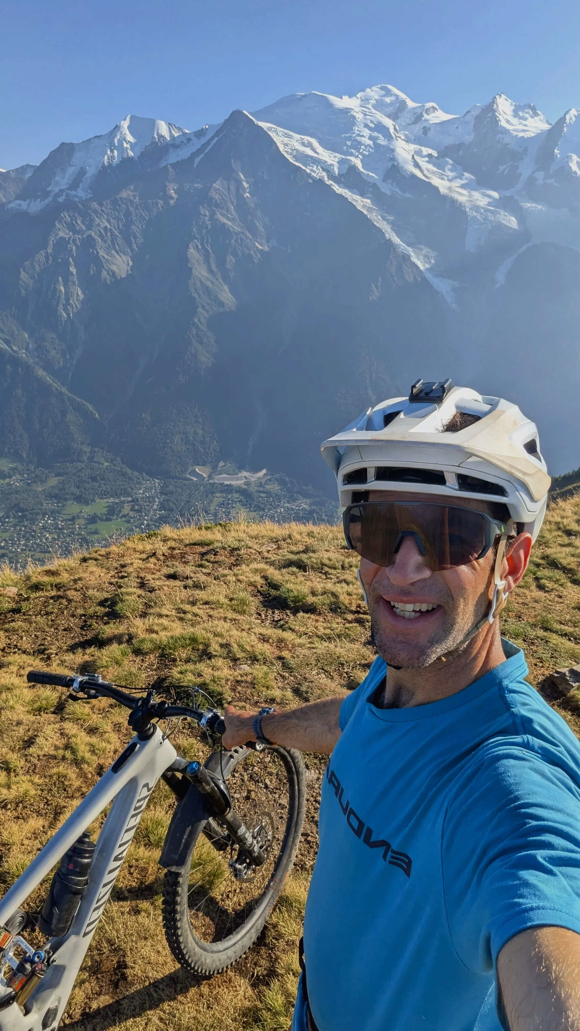 A man wearing a blue shirt, helmet, and sunglasses taking a selfie with a mountain bike, smiling against a backdrop of snow-capped mountains and a clear blue sky.