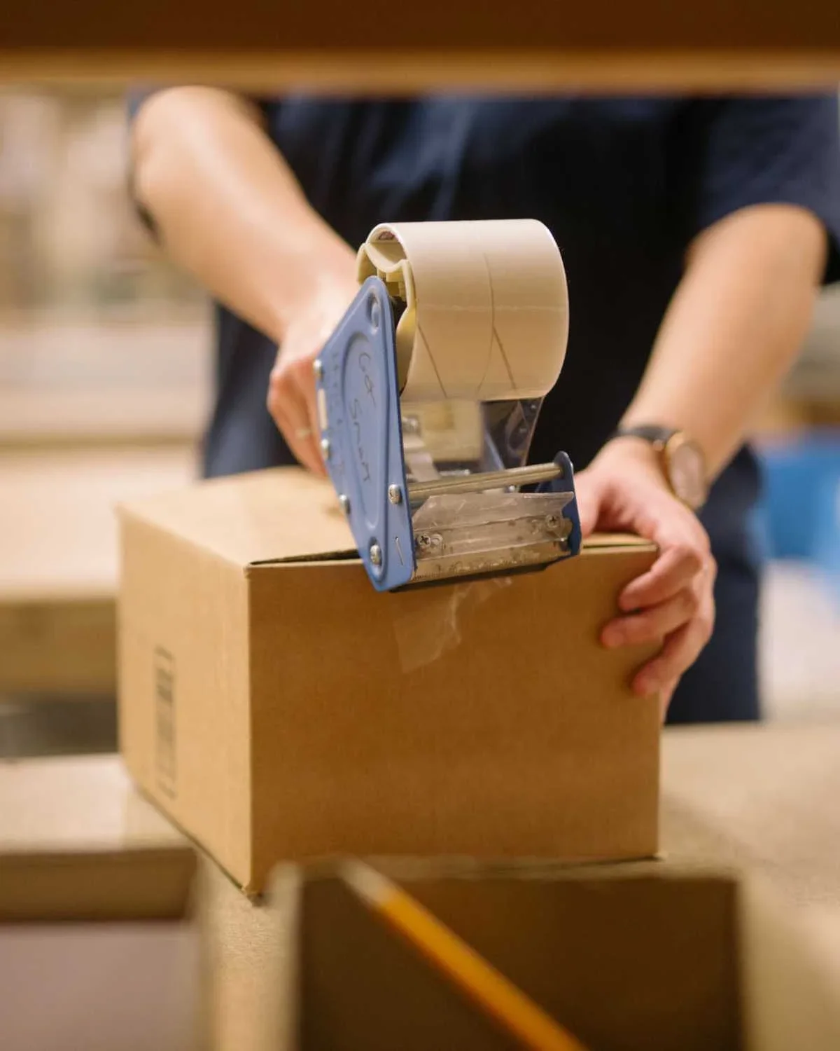Person sealing a cardboard box with clear packing tape in a warehouse or packaging setting.