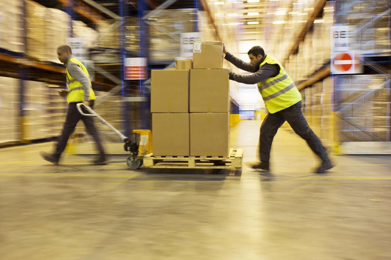 Two warehouse workers wearing yellow safety vests are moving cardboard boxes on a pallet in a large storage facility.