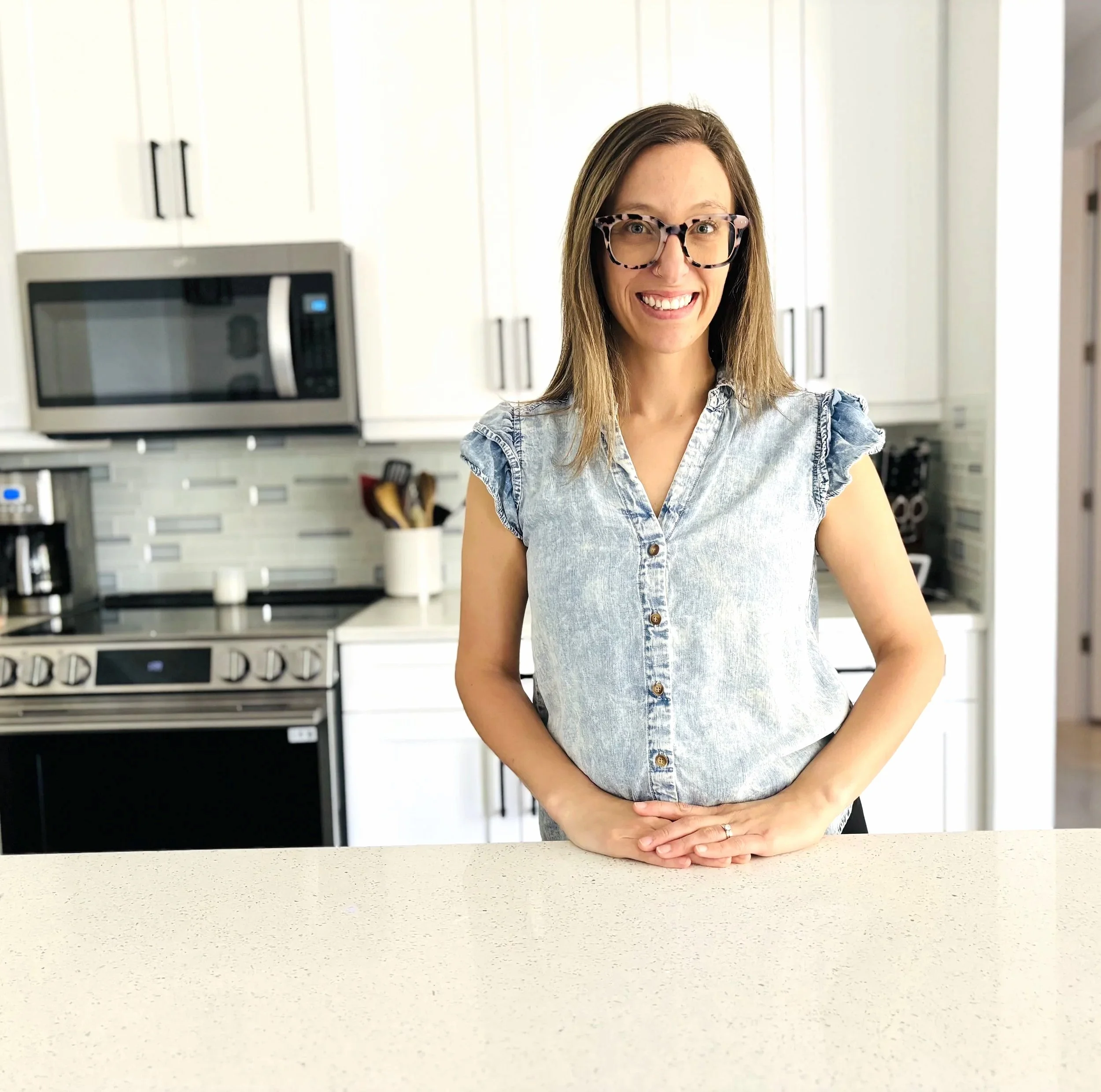 A woman with shoulder-length brown hair, wearing glasses and a sleeveless denim shirt, smiling and standing in a modern kitchen with white cabinets and stainless steel appliances.