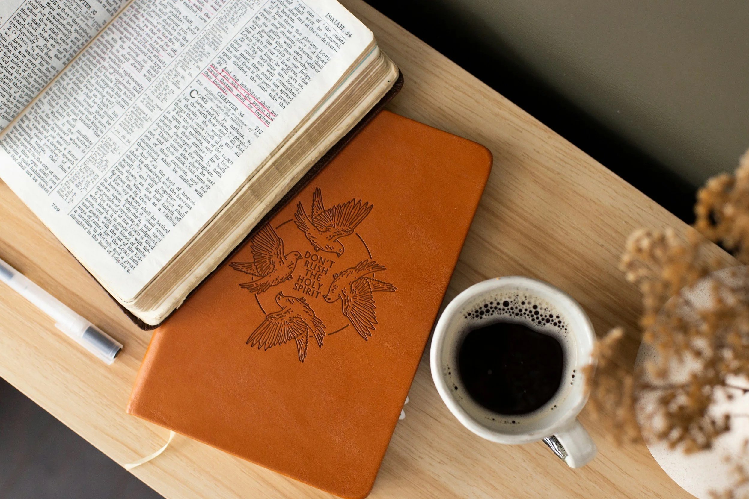 Open Bible on a wooden table next to an orange notebook with a drawing of birds and the phrase "Don't rush the holy spirit". A cup of black coffee and cookie are also on the table.