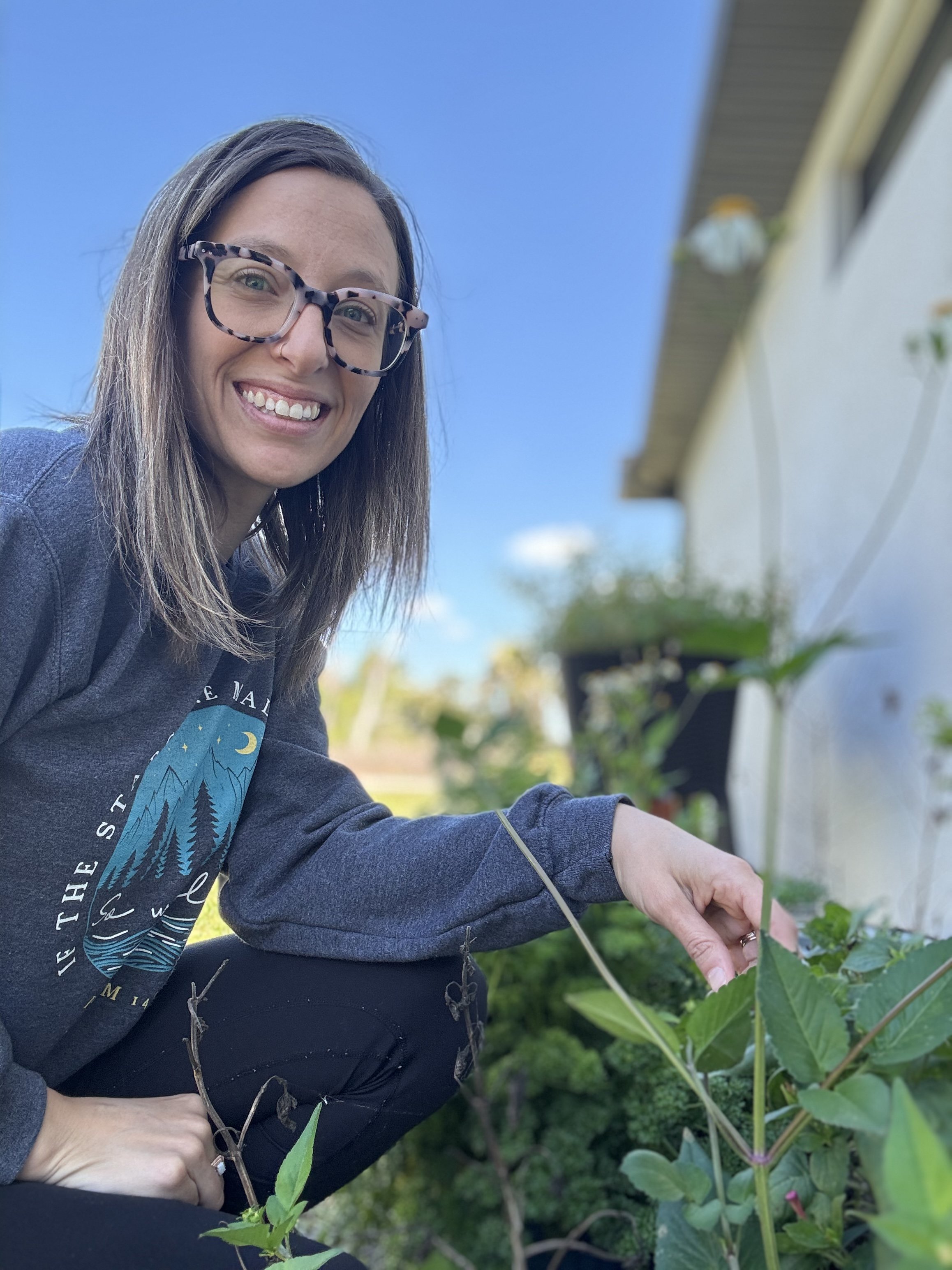 A smiling woman with glasses and shoulder-length brown hair, wearing a dark gray hoodie, crouching outdoors next to a garden wall, picking leaves or tending plants on a sunny day.