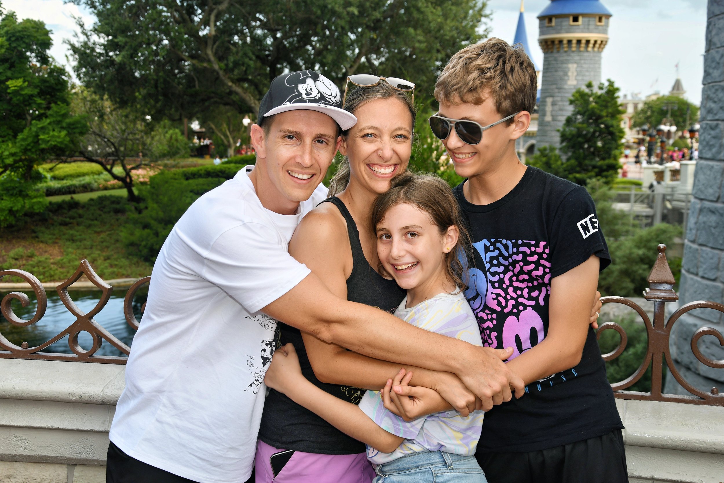 A family of four, including a father, mother, brother, and sister, hugging and smiling at a theme park with a castle in the background.