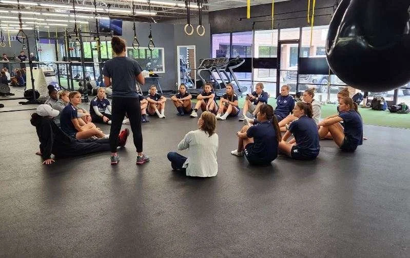 Group of women and girls sitting in a circle on the gym floor during a fitness class, with an instructor standing in the center.