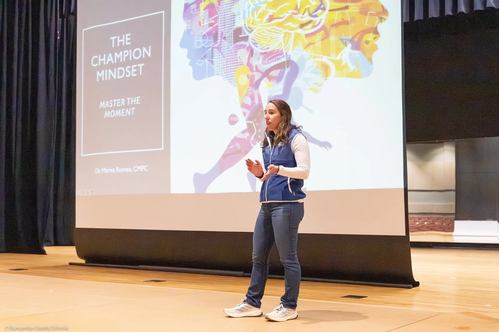 A woman giving a presentation in front of a large screen that displays a colorful graphic and text about 'The Champion Mindset,' with the subtitle 'Master the Moment,' and her name as Dr. Marisa Romeo, CMPC.