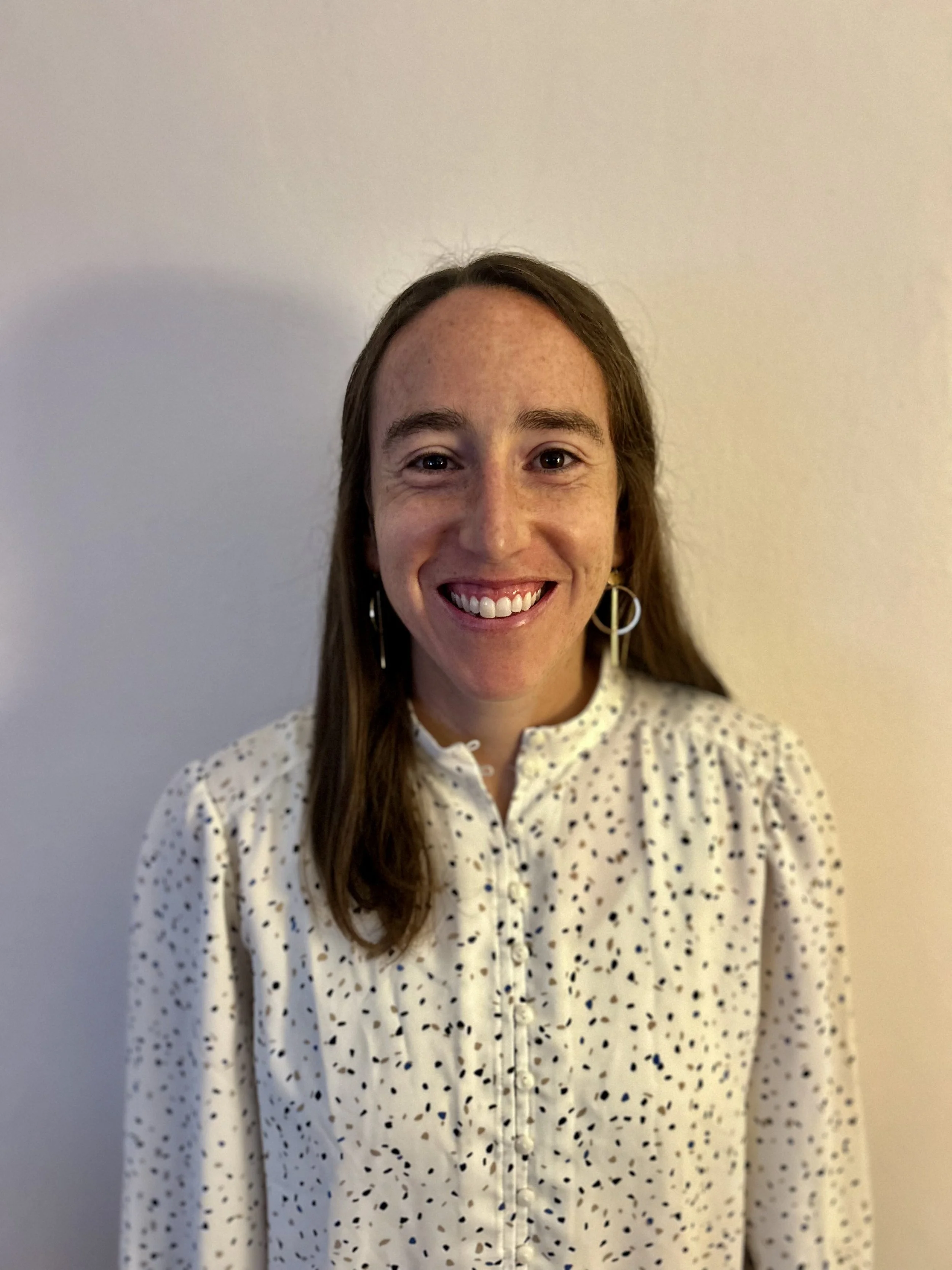 A woman with long brown hair smiling, wearing a white blouse with black spots and hoop earrings, standing against a plain light-colored wall.