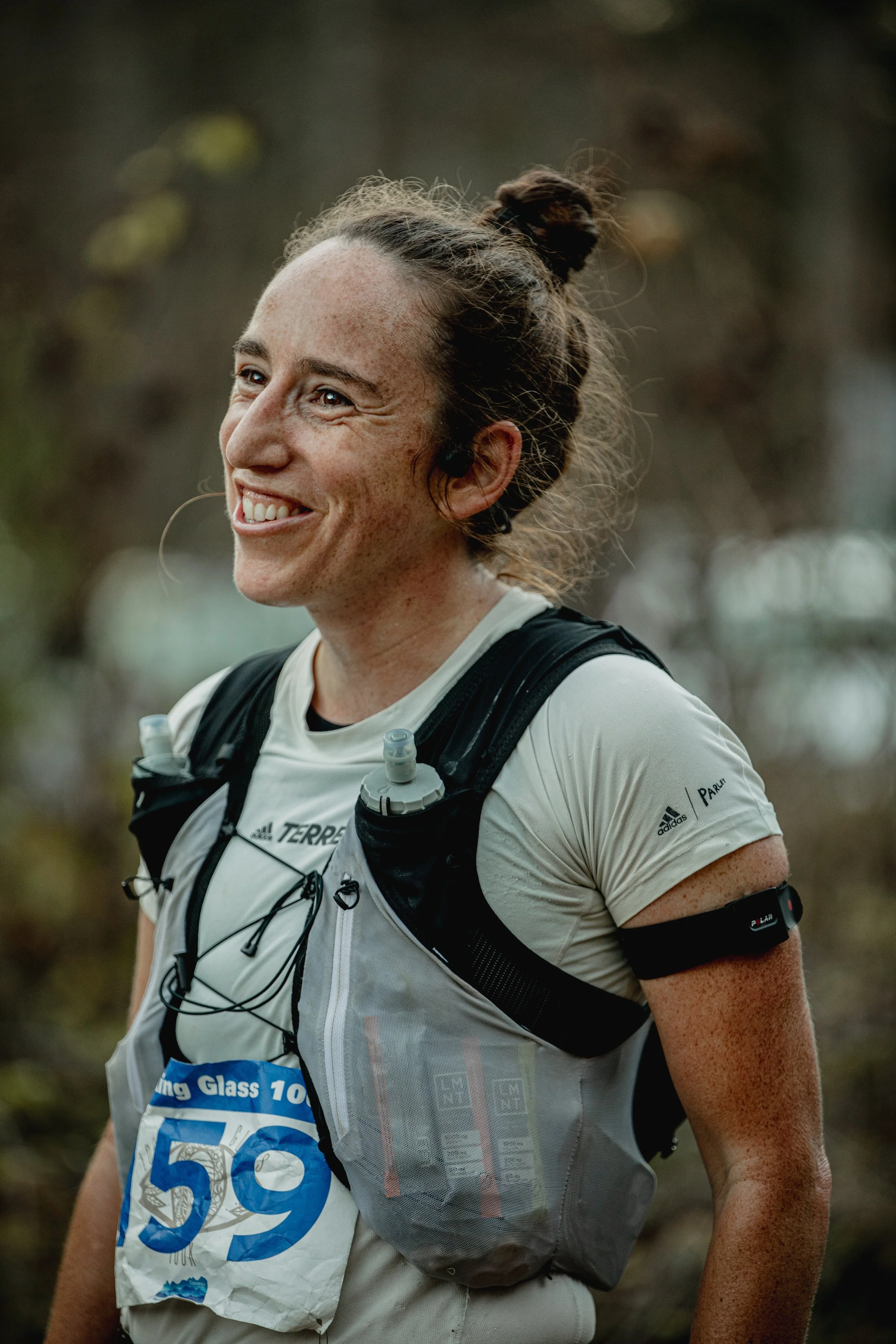 A woman with a bun hairstyle and freckles, smiling during a trail race, wearing a white athletic shirt, a running bib number 59, a hydration pack, and a fitness tracker on her arm, outdoors with blurred trees in the background.