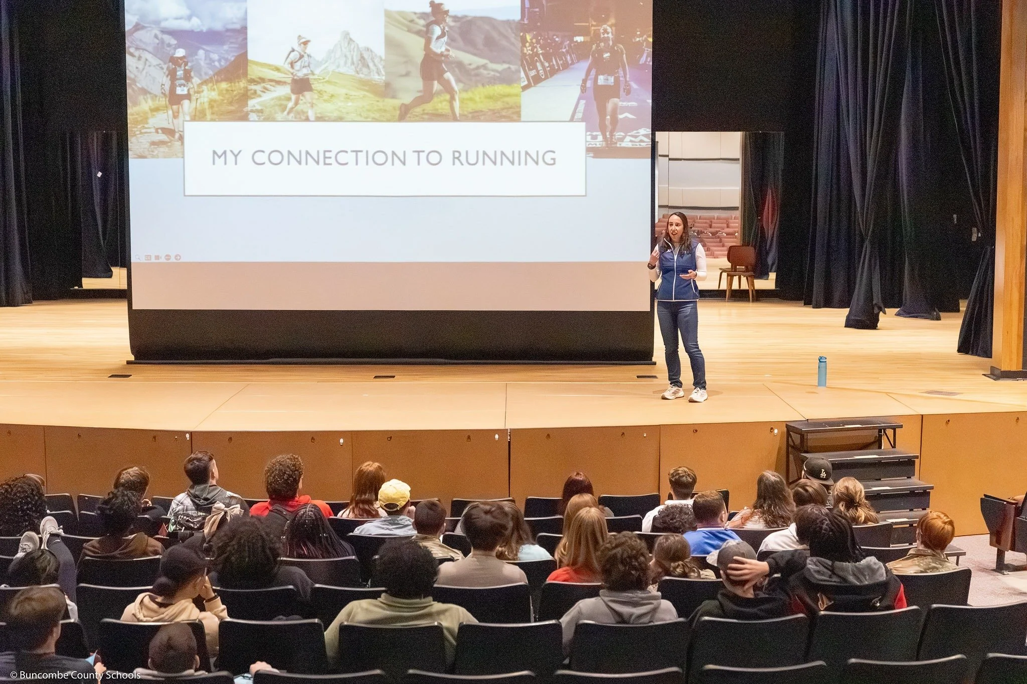A woman giving a presentation on a stage in front of a large audience. The projection screen behind her displays images of running and the text "My Connection to Running". The audience is seated and listening attentively.