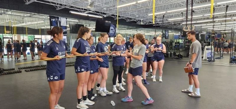 A group of female athletes in gym attire receiving instructions from coaches at a fitness center, with exercise equipment visible in the background.