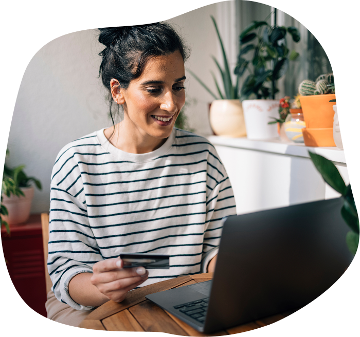 A woman with dark hair in a bun, wearing a white and black striped long-sleeve shirt, smiling while using a laptop and holding a credit card in a bright room surrounded by potted plants.