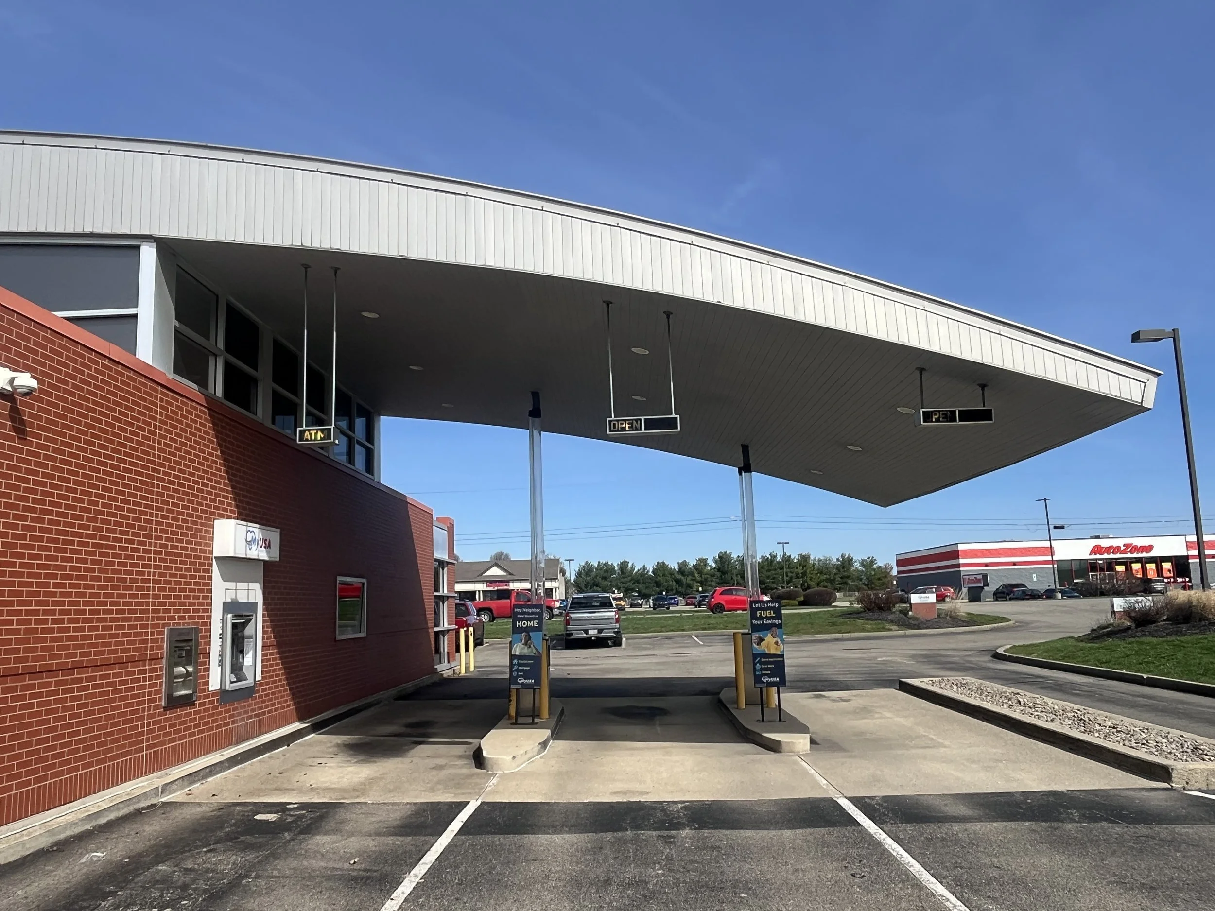 A drive-thru area of a service station with a red brick building, ATM and pay phone, and signs indicating open and pet. In the background, there is an AutoZone store and a parking lot with cars.