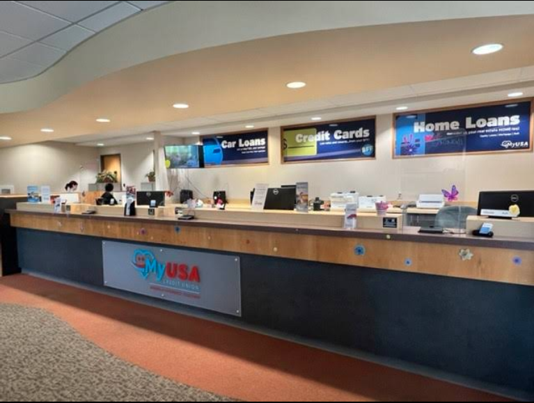 Inside a financial services office, a long counter with multiple computer stations and signs for car loans, credit cards, and home loans displayed above. The front of the counter has a sign saying 'My USA' with a patriotic design.