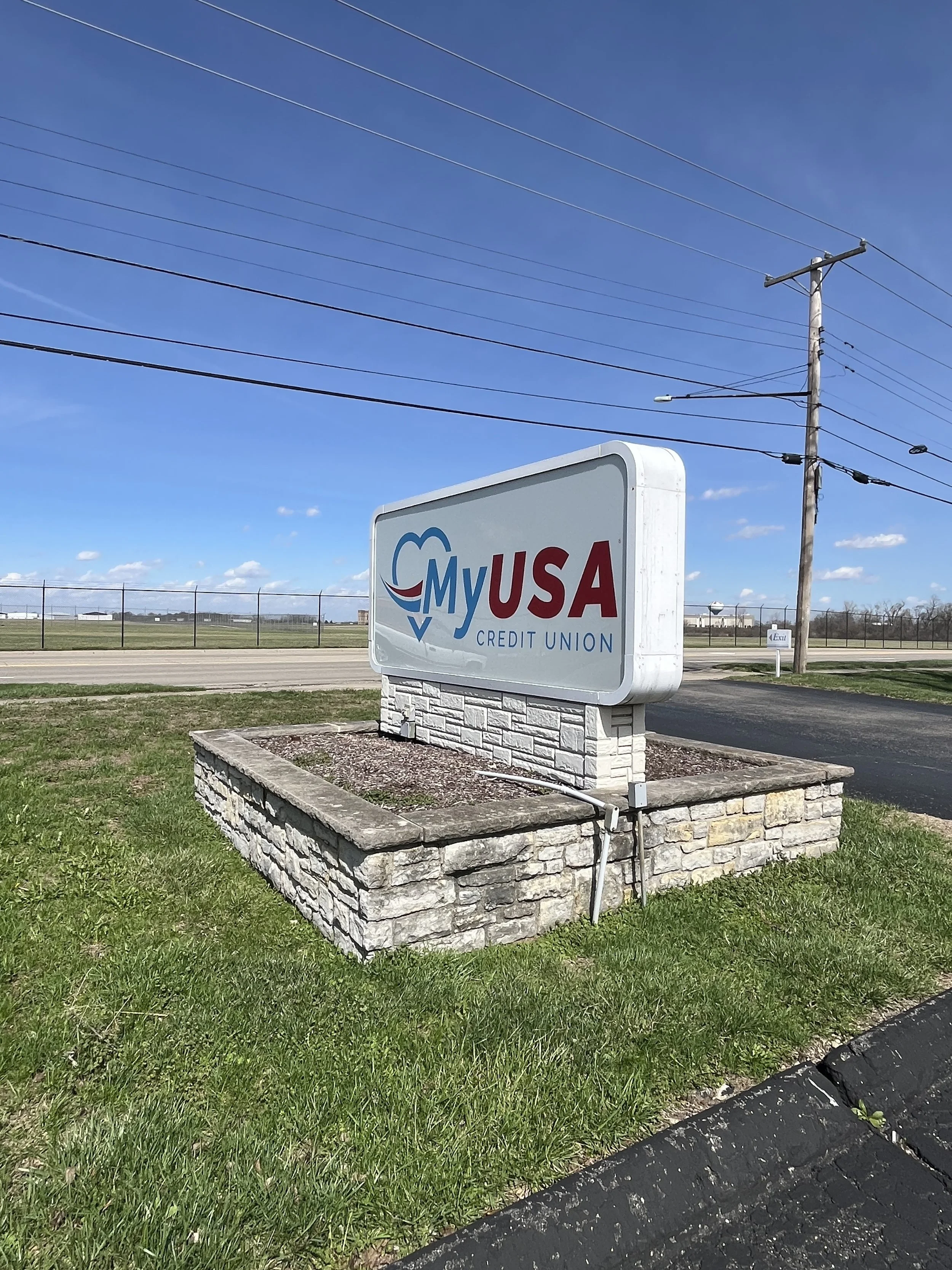 A sign for My USA Credit Union on a stone base in a grassy area, with a clear blue sky and power lines overhead.