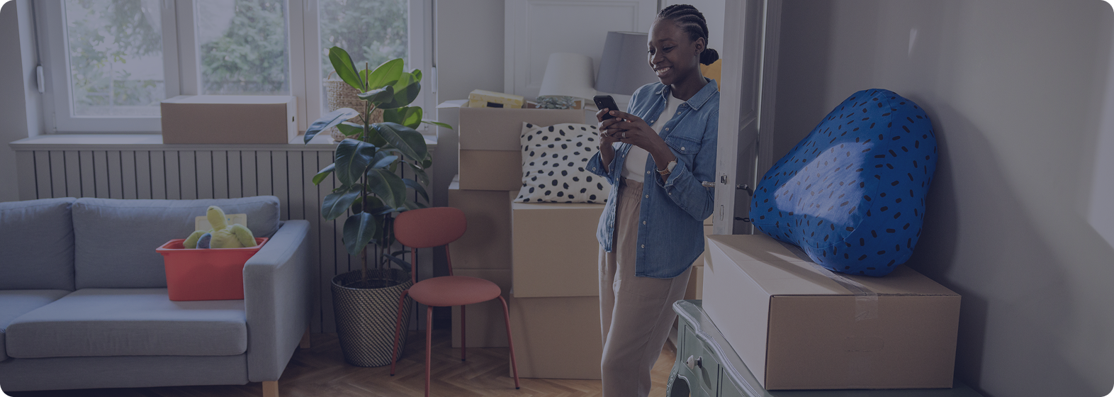 A woman standing in a living room, smiling at her phone. The room has a gray couch, a potted plant, a chair, boxes, and a large blue sleeping bag or cushion on top of a cardboard box.
