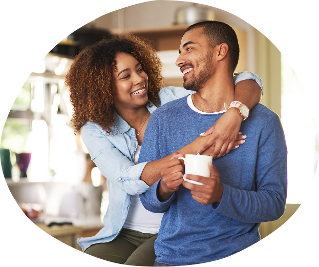 A woman wraps her arms around a man holding a white mug, both smiling joyfully in a cozy kitchen setting.