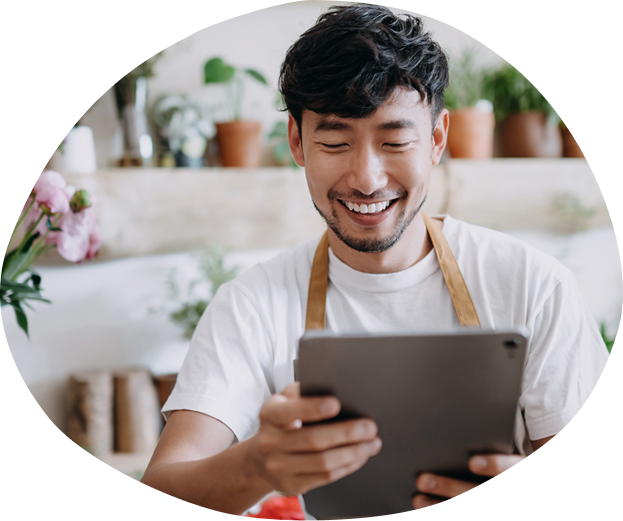 Smiling man in a white shirt and apron looking at a tablet in a cozy, plant-filled room.