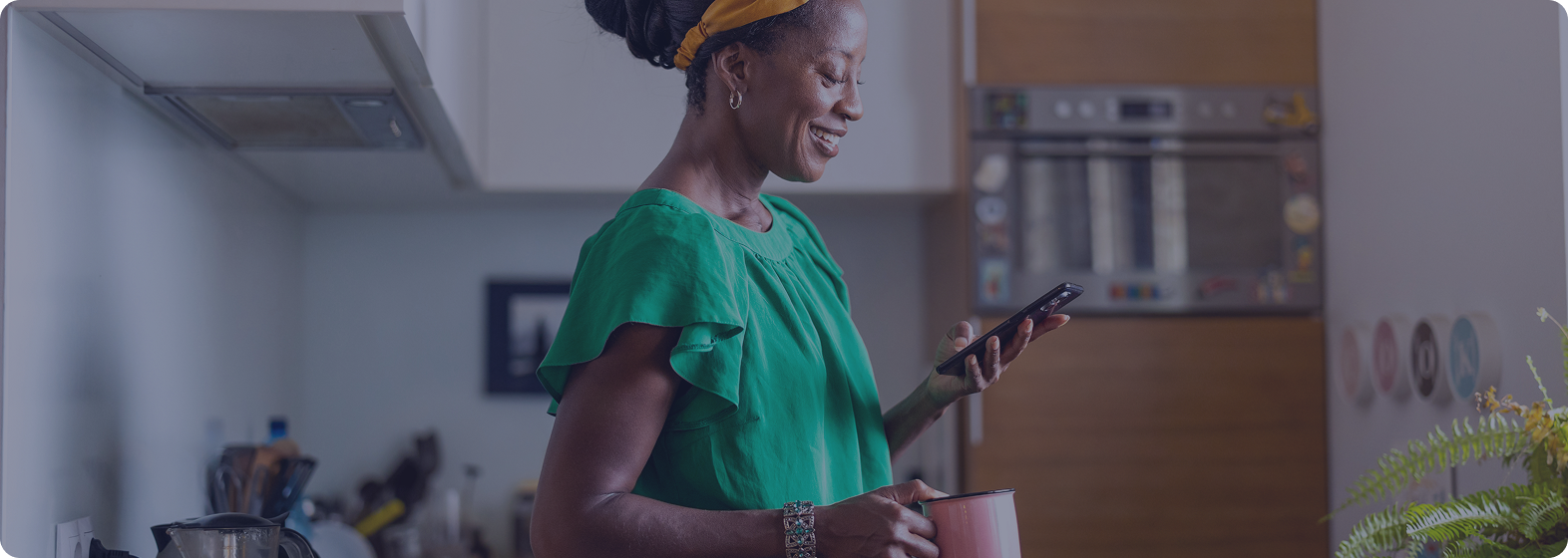 A woman with dark hair and a yellow headband, wearing a green blouse, stands in a modern kitchen smiling while looking at her phone. She holds a pink mug in her right hand.