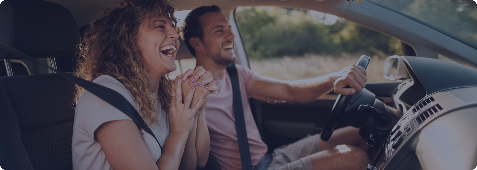 A smiling woman and man enjoying a road trip inside a car, with the woman in the passenger seat and the man driving.