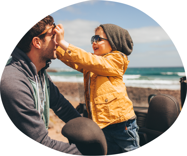 A father and young child at the beach, with the child playfully touching the father's face. The child is wearing a gray beanie, sunglasses, and a yellow jacket, while the father is dressed casually in a hoodie and jacket. The background shows ocean waves and a partly cloudy sky.