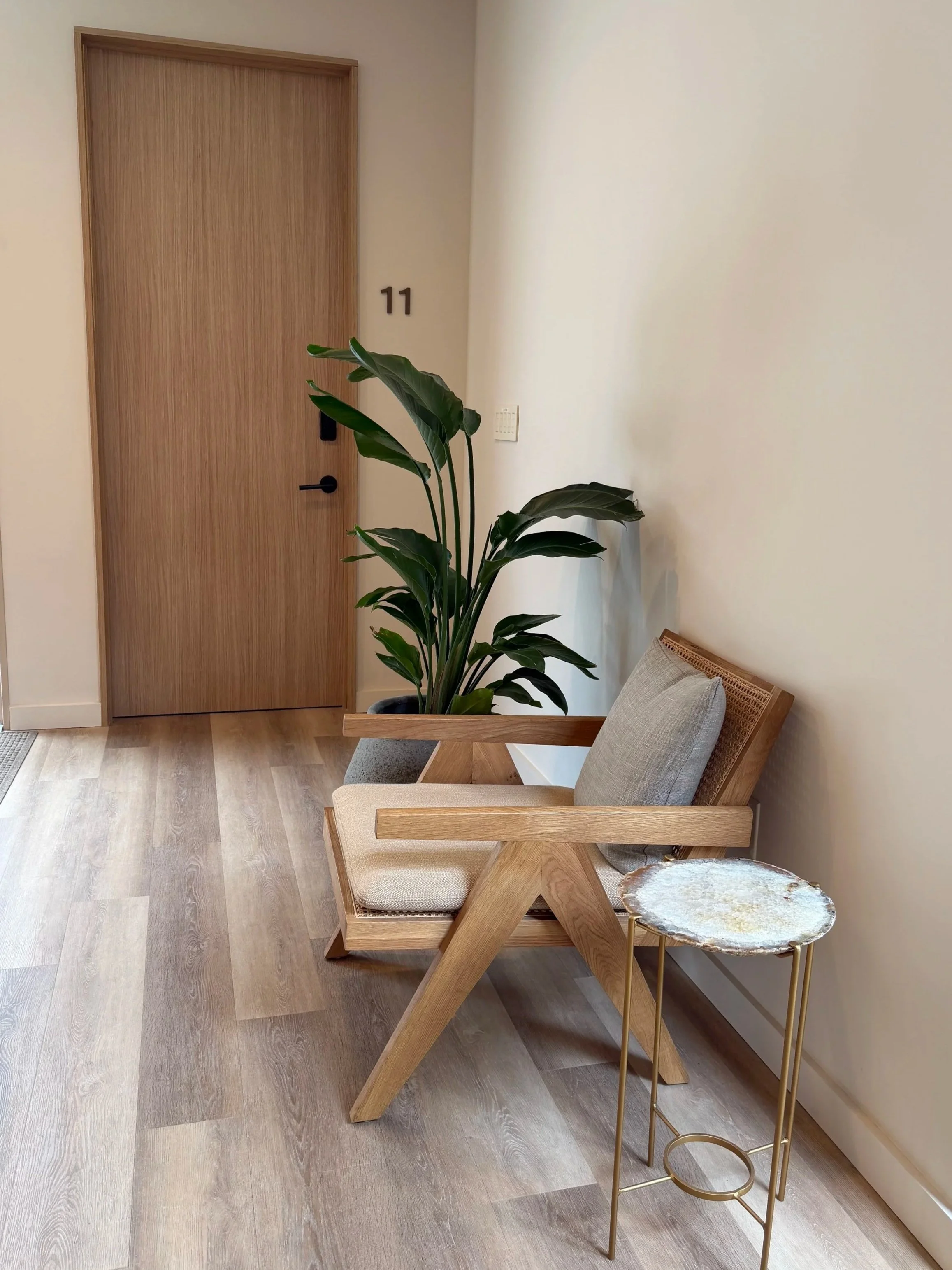 A wooden armchair with beige cushions, a gold side table with a white and gold decorative top, a large house plant in a black pot, and a wooden door marked with the number 11. The room has a light-colored wall and wood flooring.