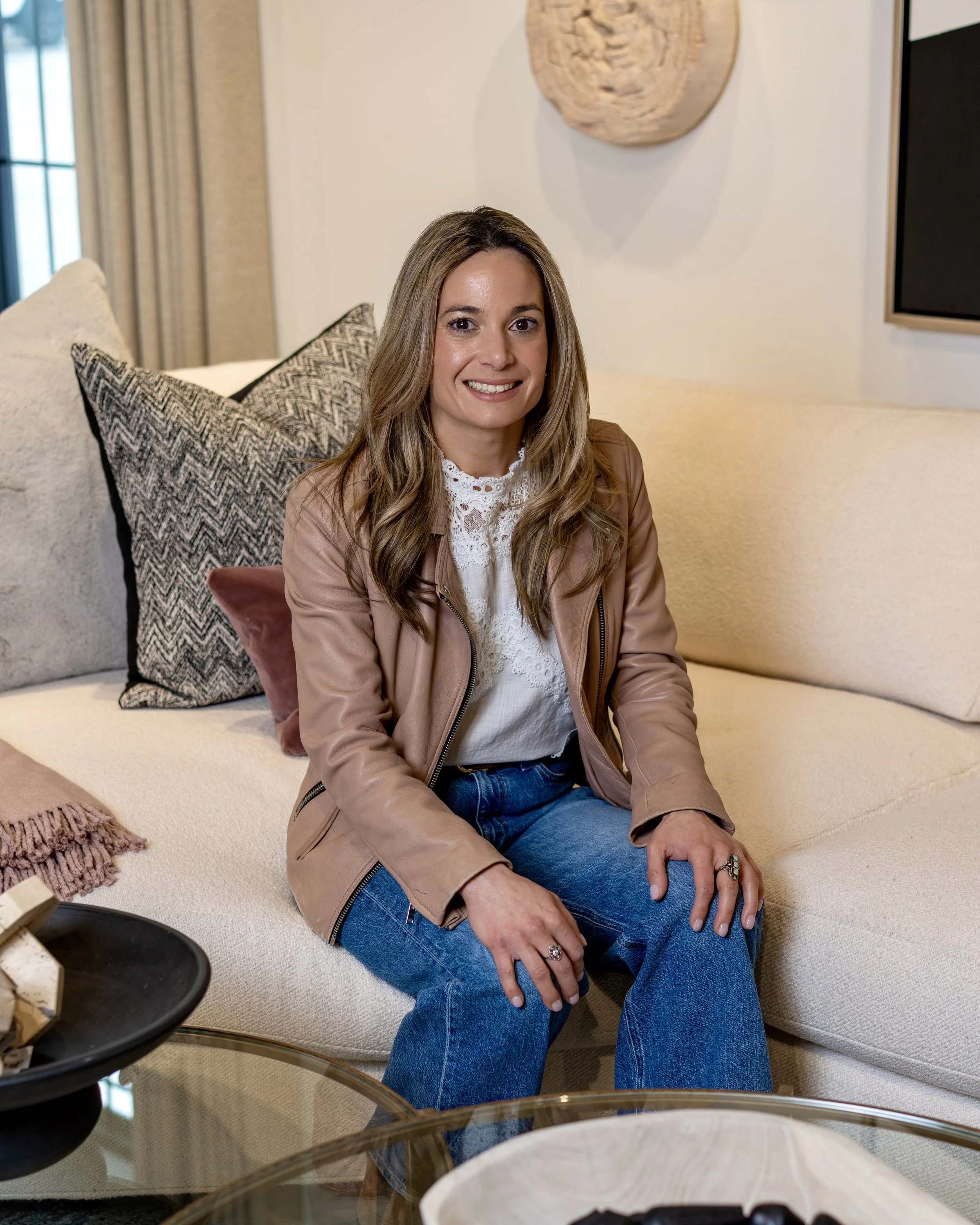 A woman with long wavy hair, wearing a light brown leather jacket, white lace top, and blue jeans, sits on a cream-colored sofa with textured pillows. She is smiling and looking at the camera, in a cozy, well-decorated living room with abstract wall art.