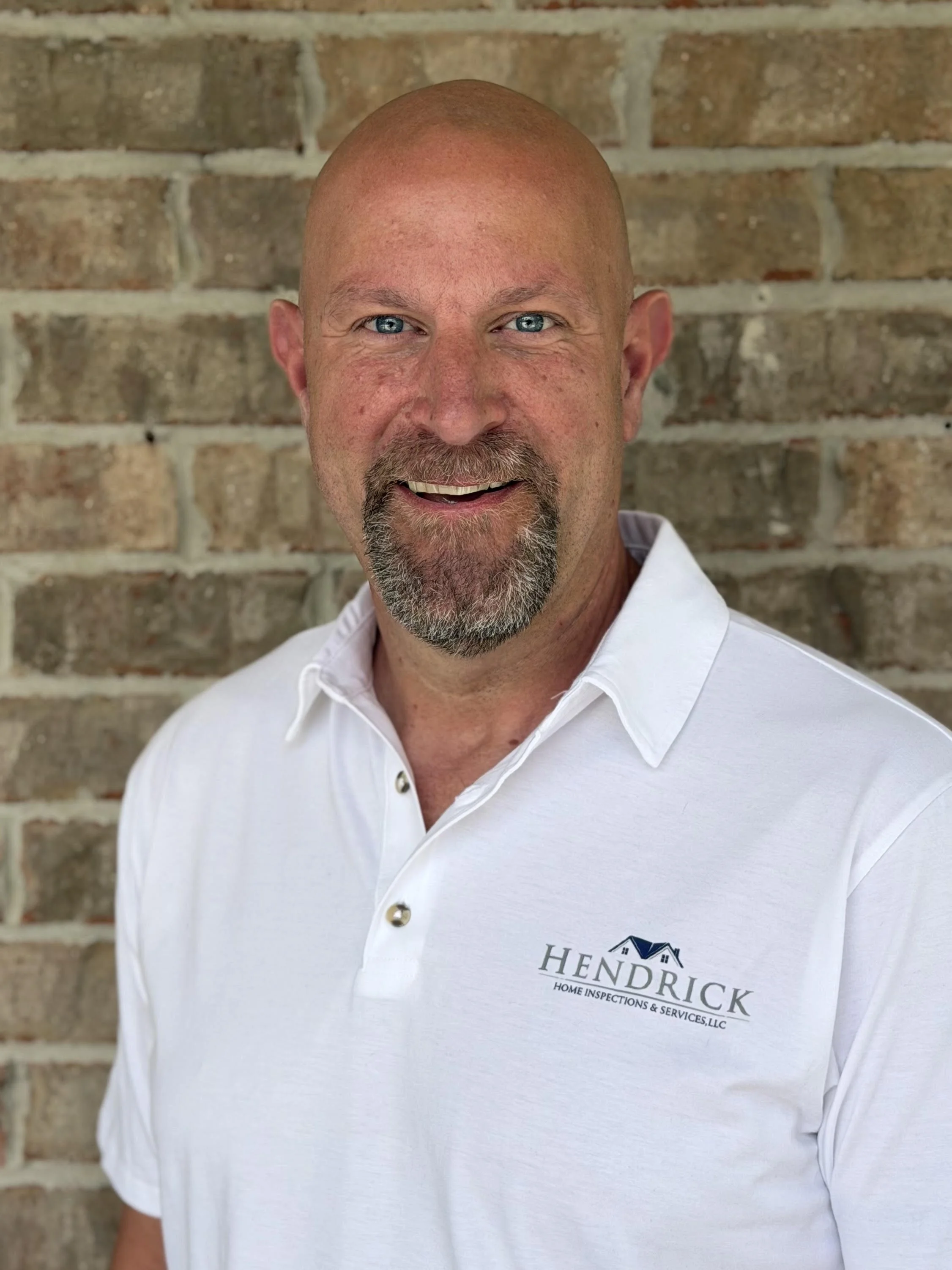 A man with a shaved head and beard, wearing a white polo shirt with a company logo, standing in front of a brick wall.