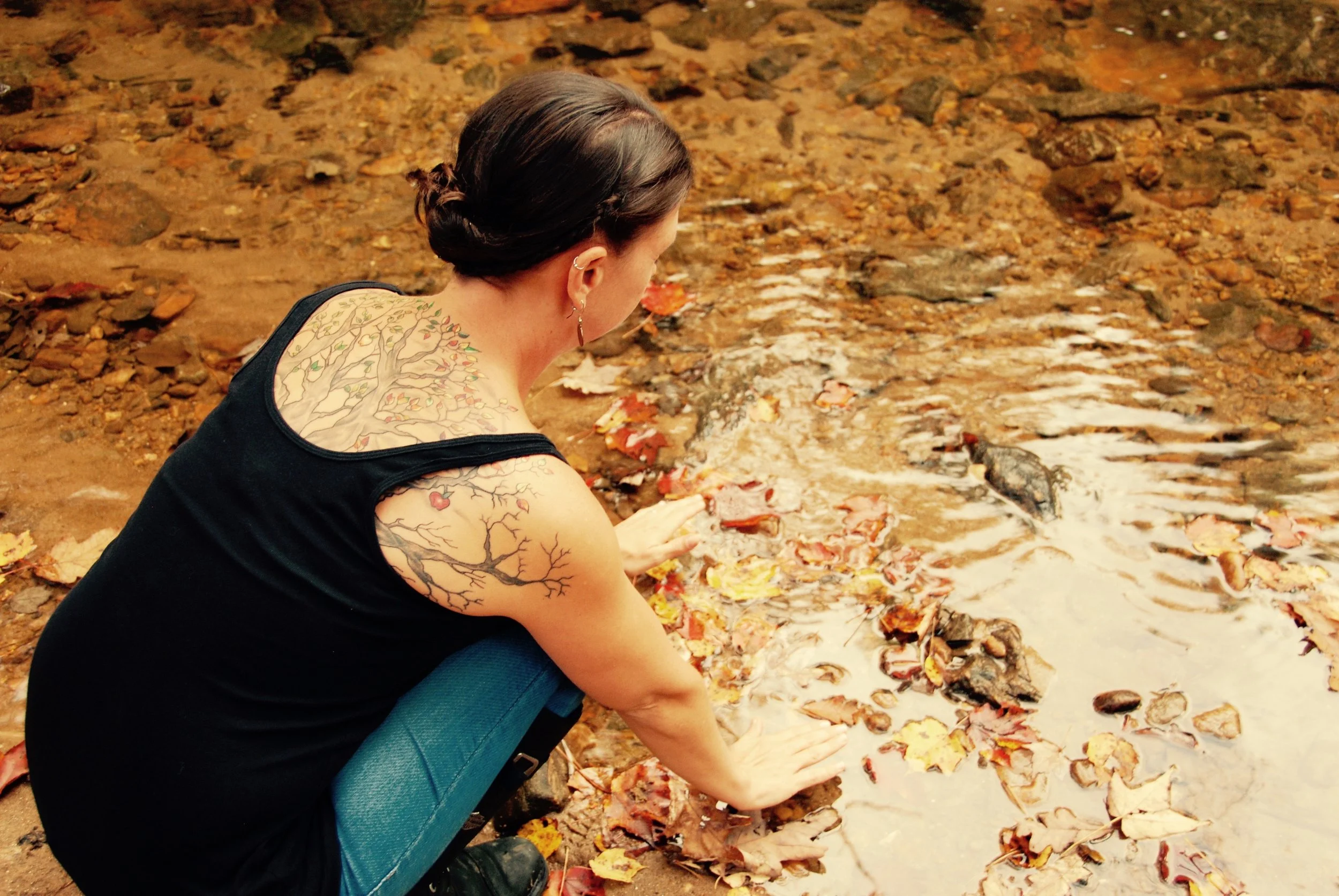 A woman with tattoos on her shoulders and arm, wearing a black top and blue jeans, sitting by a shallow stream, touching the water amid fallen autumn leaves.
