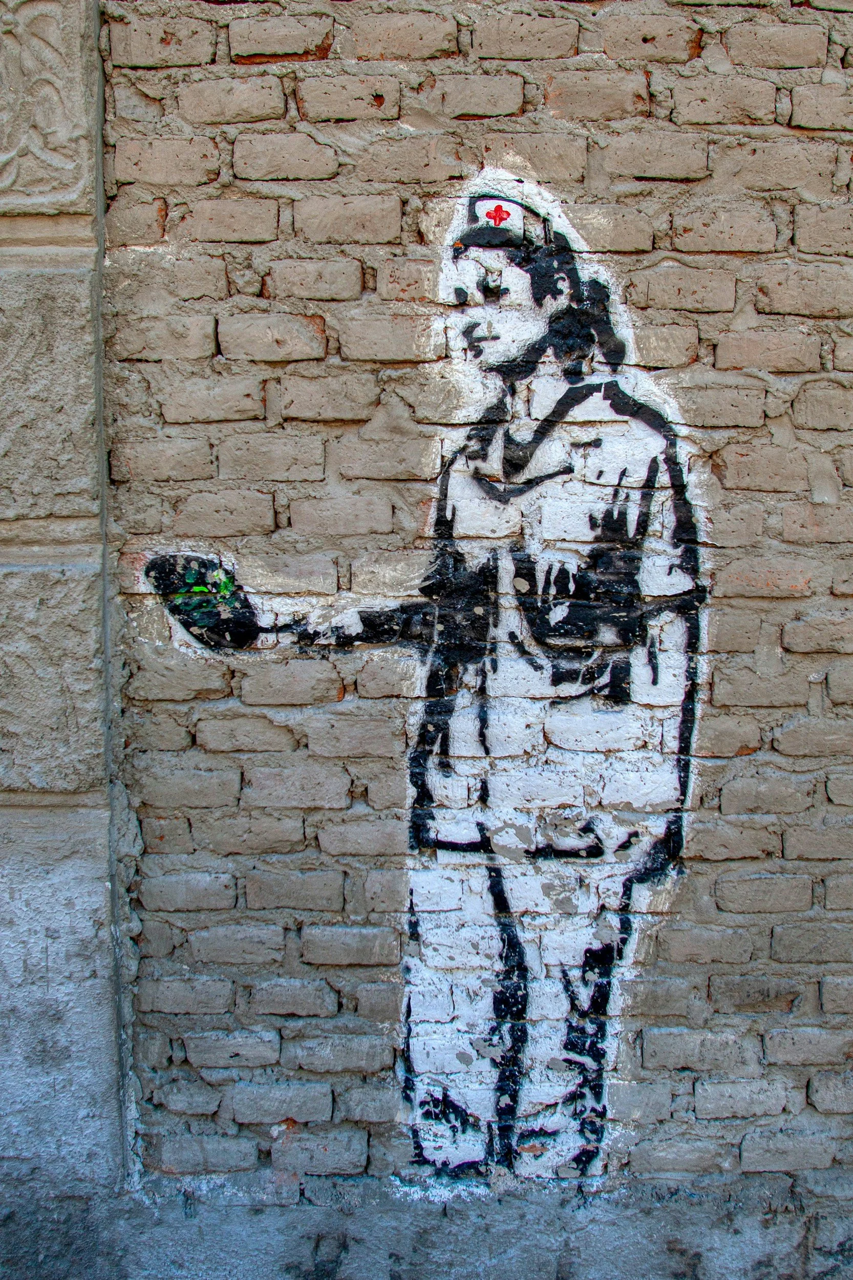 Street art graffiti of a nurse in a white uniform with a red cross nurse cap, holding a small green object, painted on a brick wall.