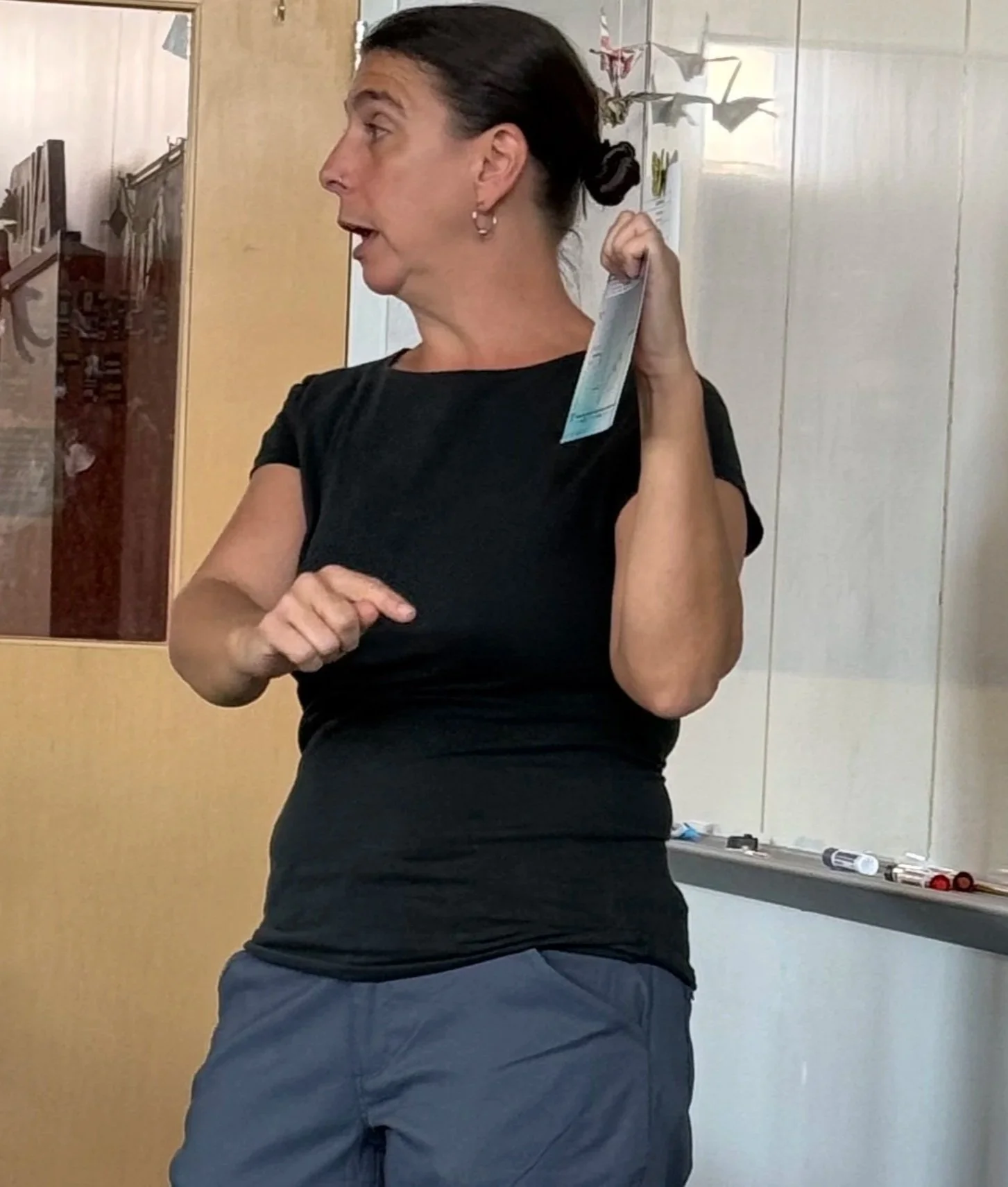 Woman with dark hair tied back, wearing a black t-shirt and gray pants, standing in a room with a whiteboard and some markers behind her, holding a face mask in her right hand.