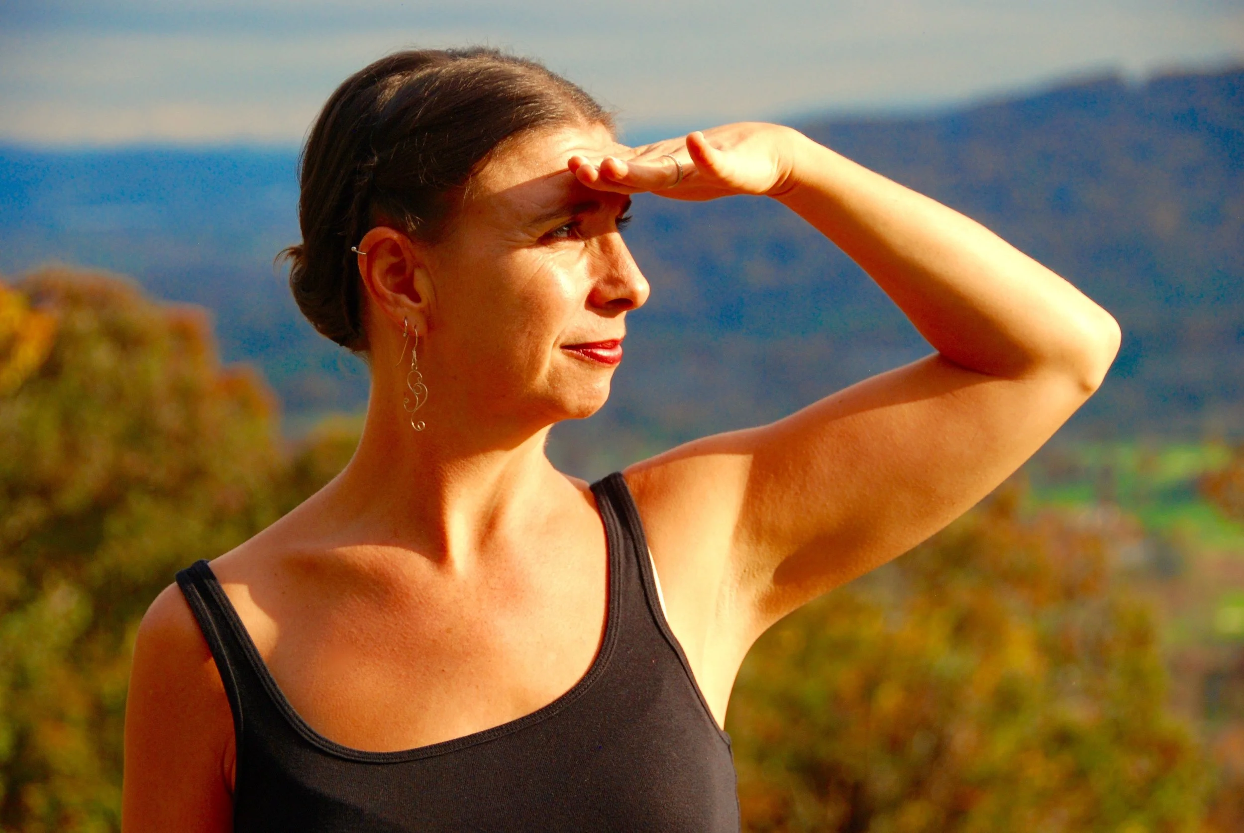 Woman shielding her eyes from the sun outdoors with nature scenery in the background.