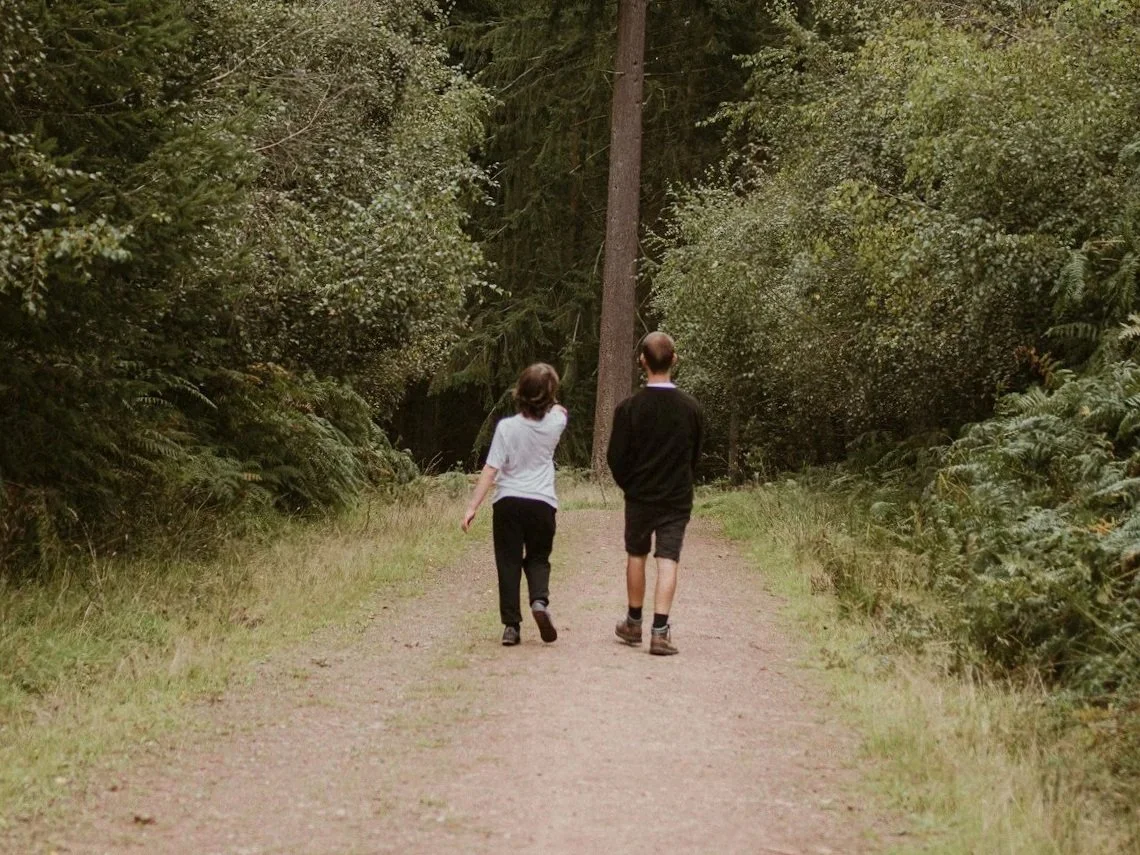 Two people walking on a dirt trail through a lush green forest.