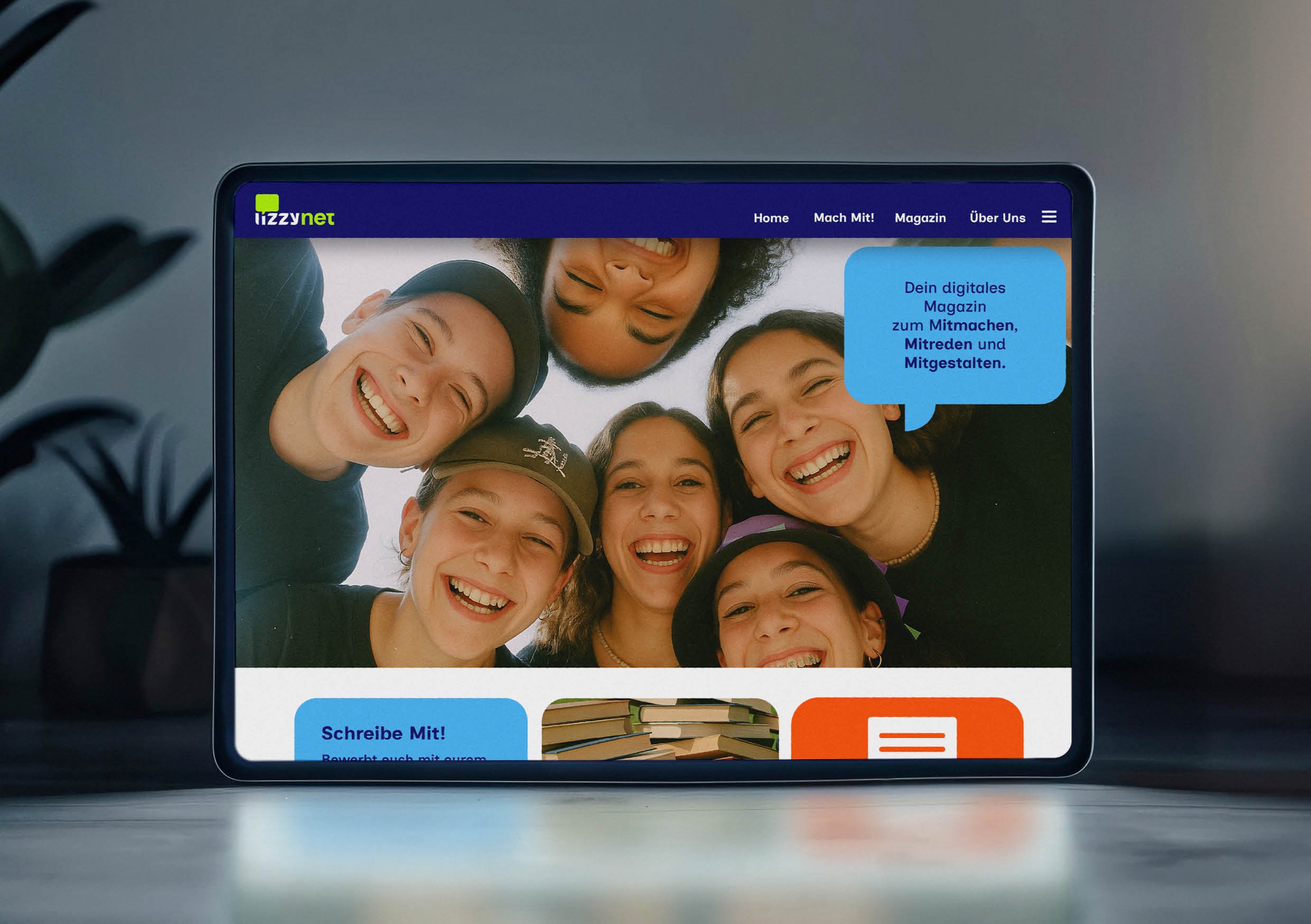 Ein Tablet zeigt eine Webseite mit einem Gruppenfoto lachender junger Menschen und einem blauen Sprechblasen-Text, der zu digitaler Mitgestaltung aufruft.