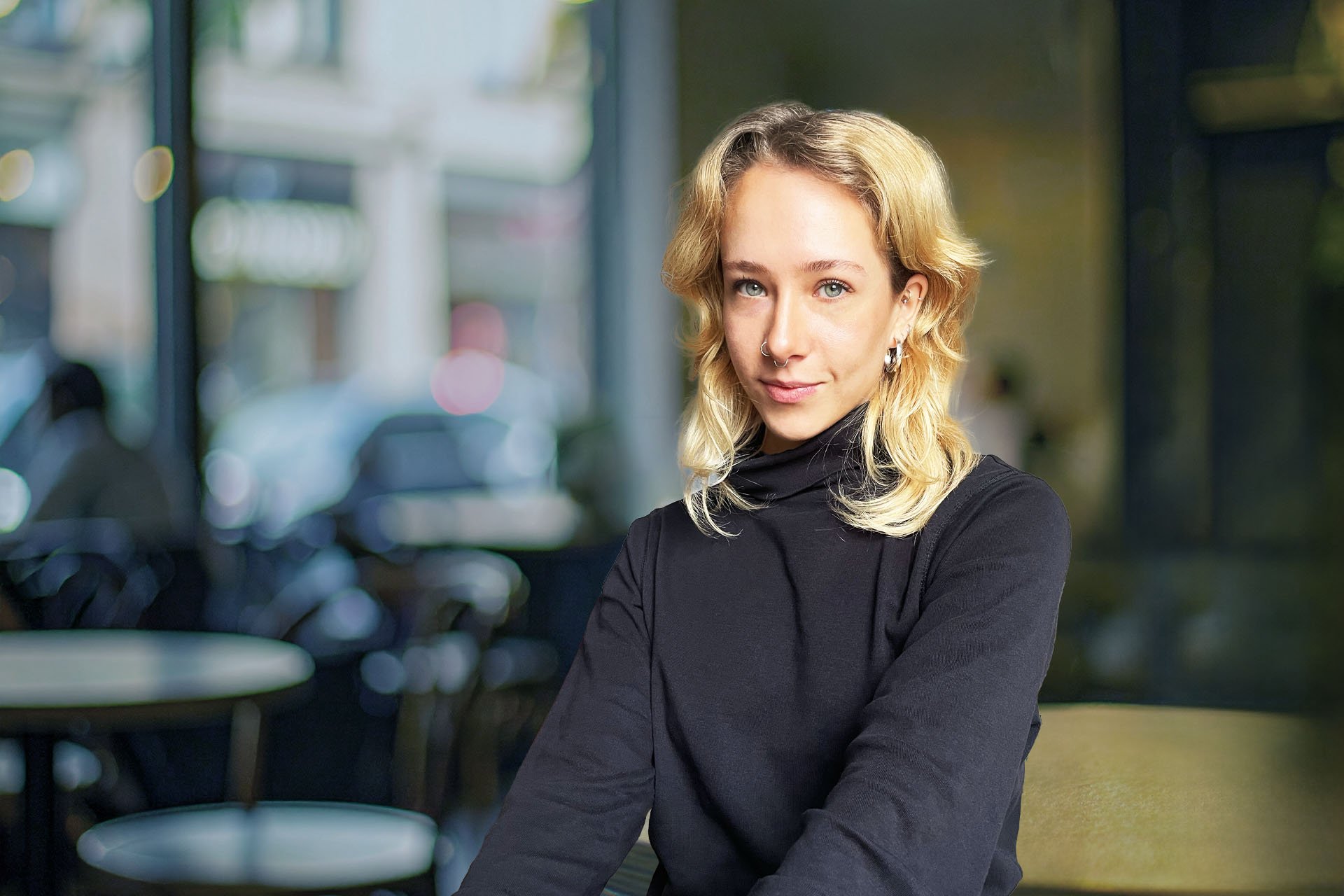 Junge Frau mit blondem, gewelltem Haar, Piercings, dunkler Kleidung und einer selbstbewussten Haltung sitzt in einem Café.