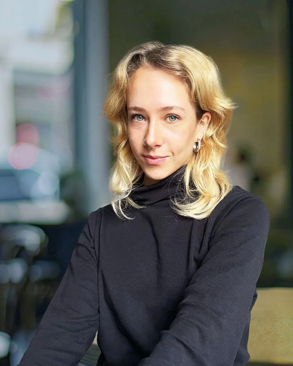 Junge Frau mit blondem, gelocktem Haar, schwarzen Pullover und Ohrringen, steht vor einem unscharfen Hintergrund in einem modernen Café oder Restaurant.