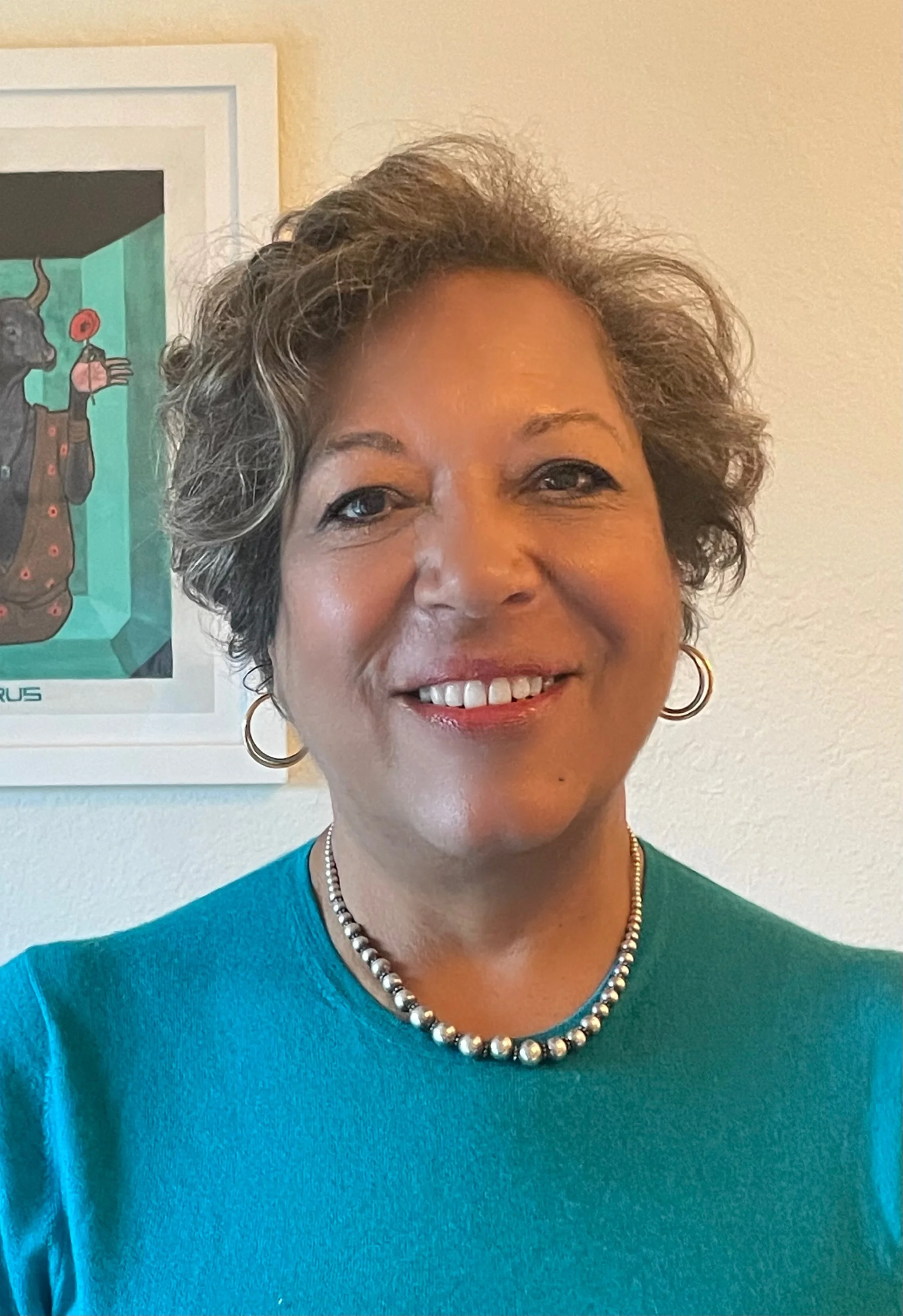 Smiling woman with short, curly gray hair wearing a turquoise top, silver beads, and hoop earrings, standing in front of a colorful framed art piece on a white wall.