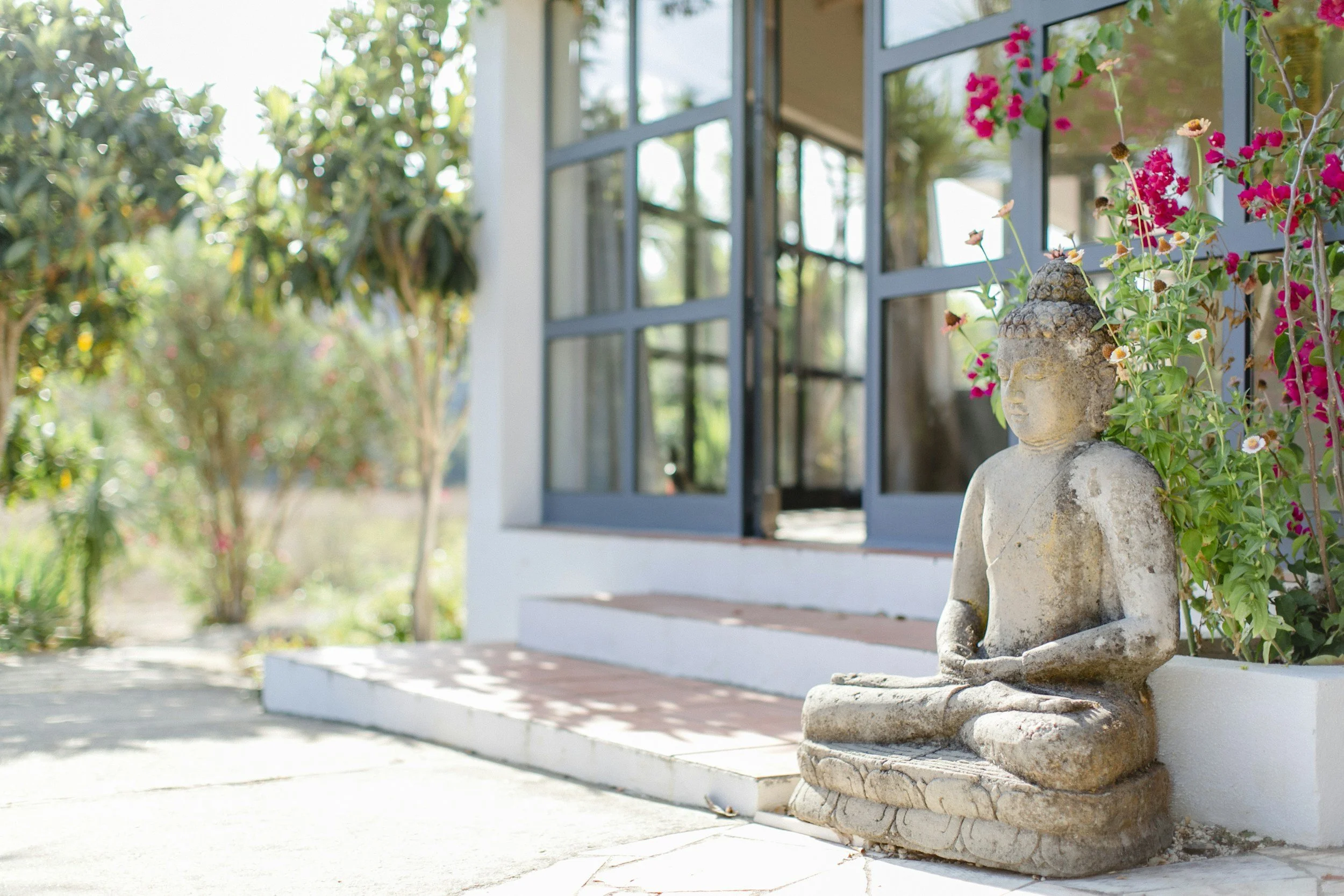 A stone Buddha statue is seated outdoors near a white building with open windows and pink flowers growing beside it. The background shows green trees and a bright, sunny day.