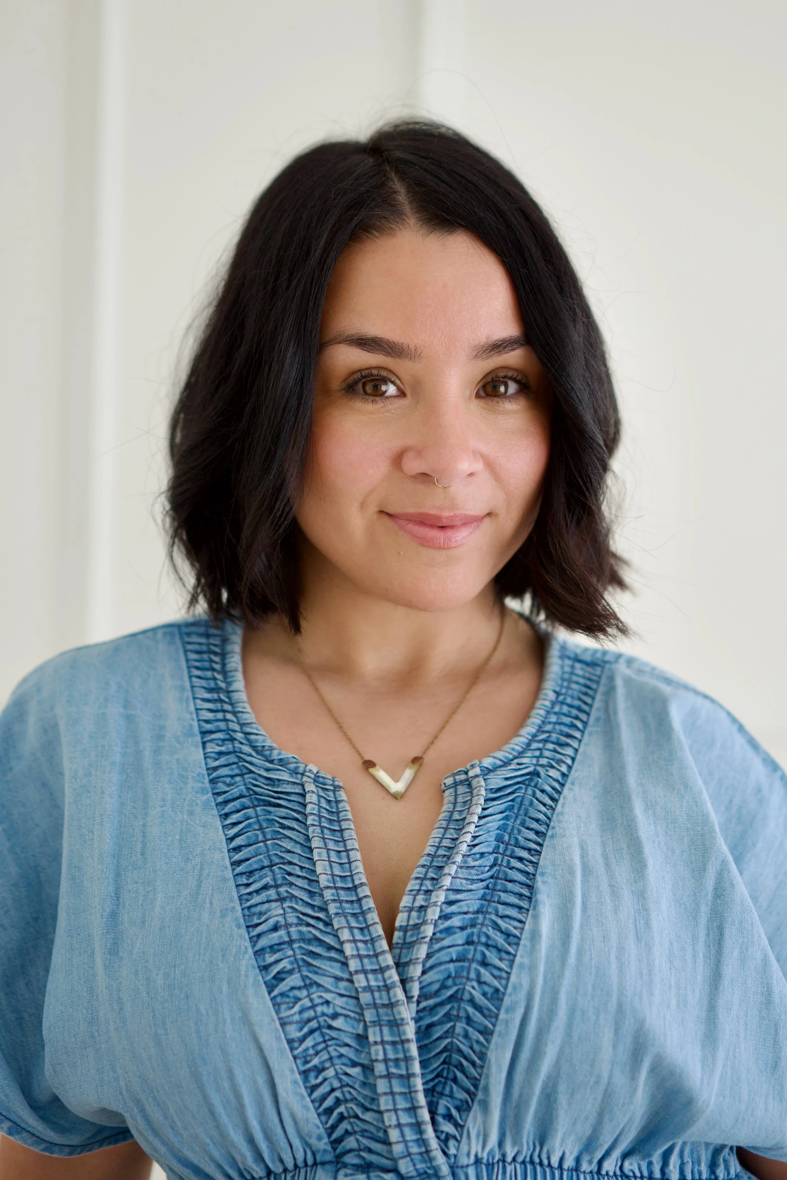 Meredith Cashman, therapist in Angora Hills, with brown hair, light skin, and brown eyes, smiling slightly in front of a plain light-colored wall. She wears a blue top with a decorative front and a gold necklace with a geometric V-shaped pendant.
