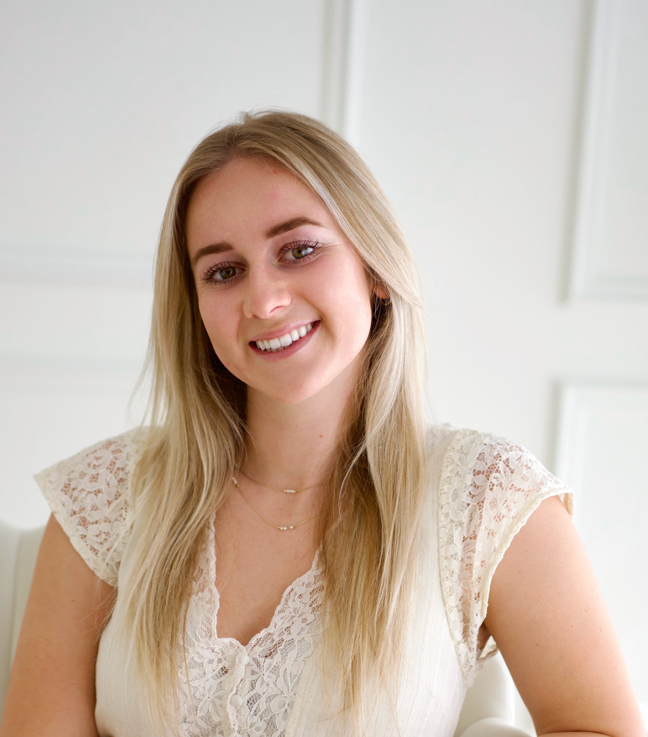 Paige Lewis, administrative coordinator at Soulfire Psychotherapy, with long blonde hair wearing a white lace top and gold jewelry, sitting indoors with a white background.