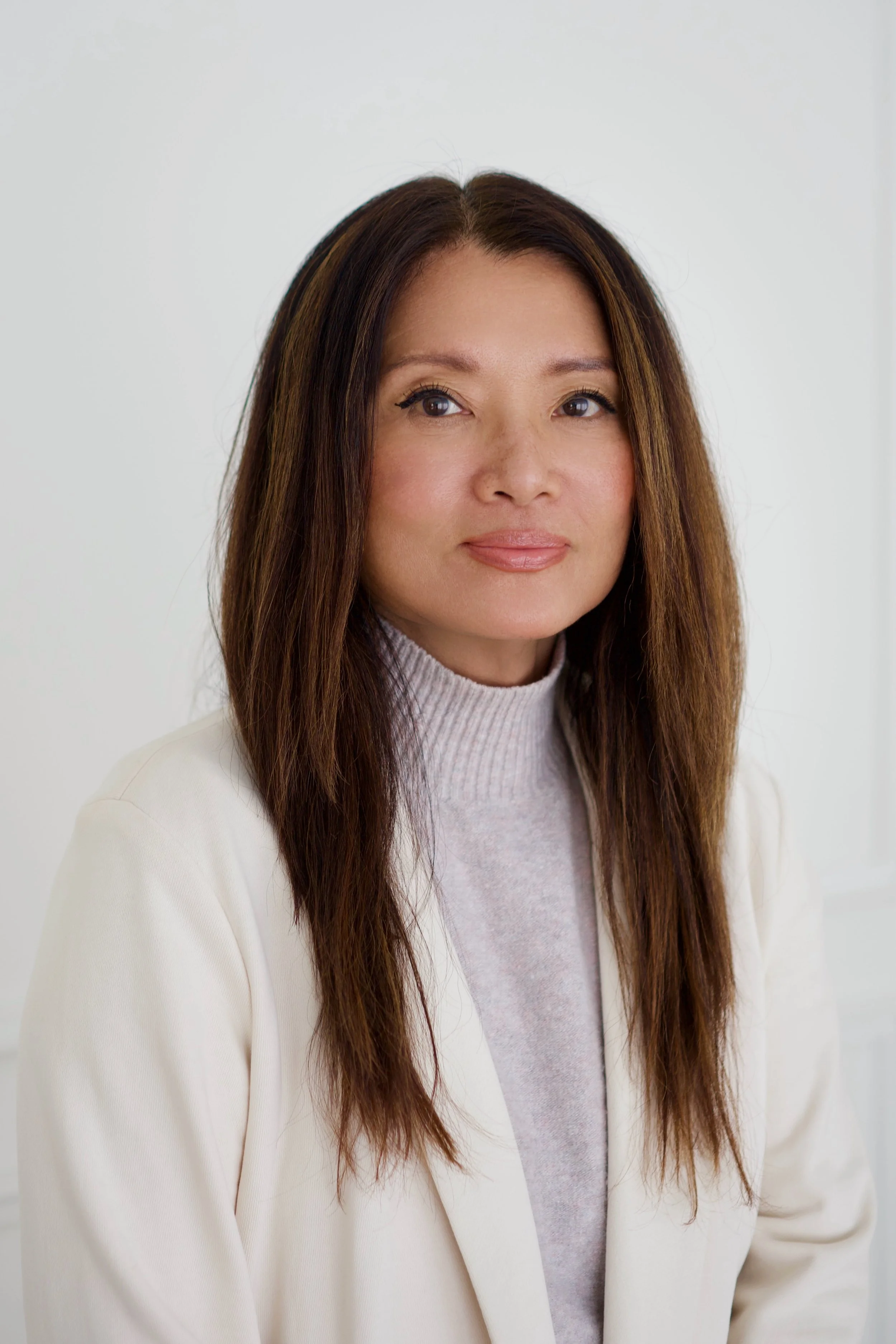 Caroline Kim, associate therapist in Angora Hills, with long brown hair, wearing a white blazer and a light-colored turtleneck sweater, standing against a plain white background.