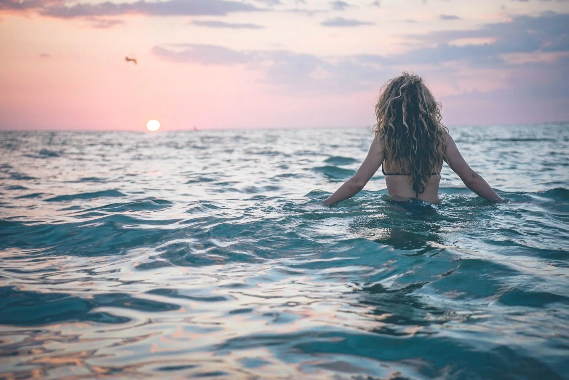 A woman with long curly hair in a swimsuit standing in the ocean at sunset, facing away from the camera, with a small boat and flying bird in the sky.
