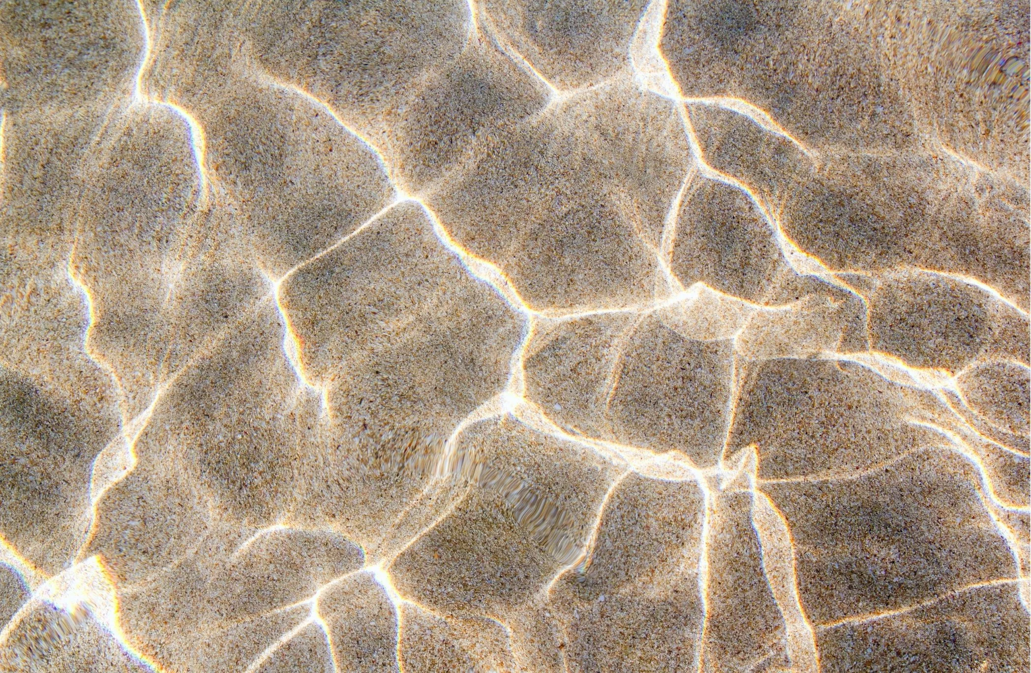 Close-up of sand and water on a beach with sunlight creating rippling patterns on the surface.