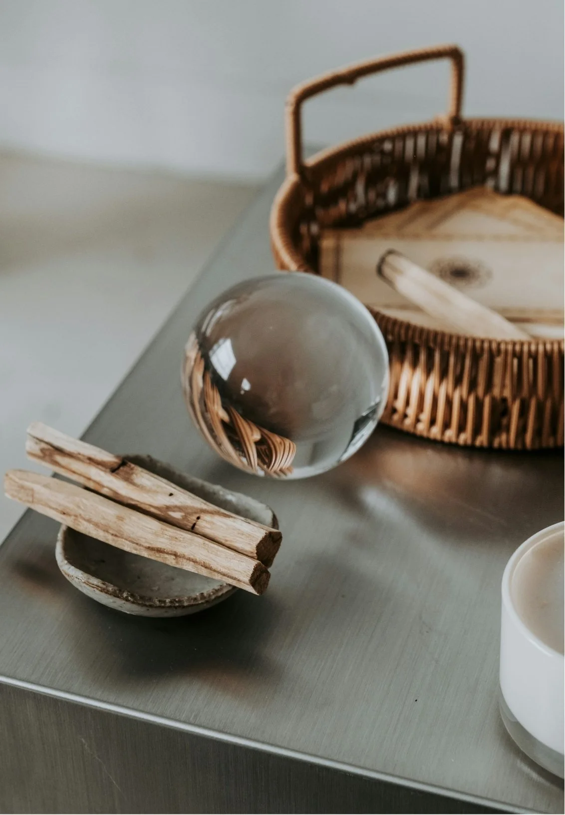 A glass sphere reflecting a wicker basket, a wooden box, and part of a table. Next to the sphere are two small wooden sticks resting on a ceramic plate. The items are on a gray surface.