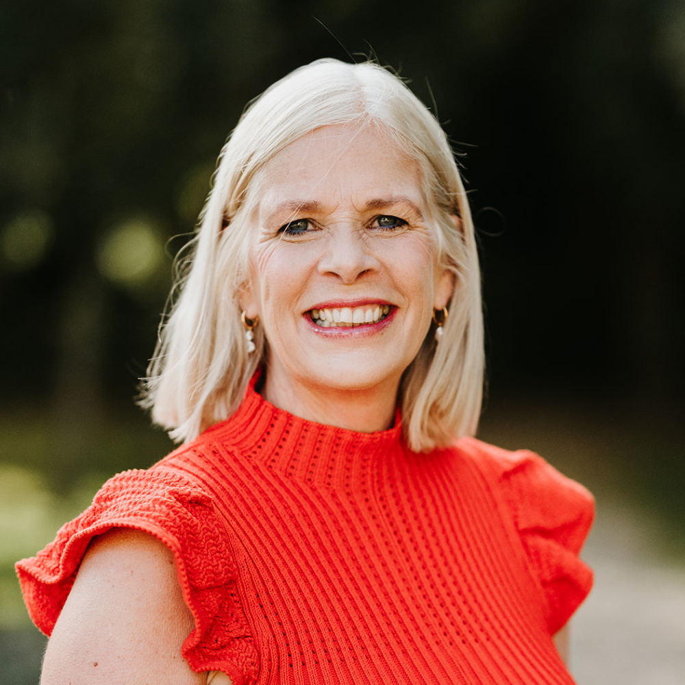A smiling woman with shoulder-length blonde hair, wearing a red textured sleeveless top, standing outdoors with a blurred green background.