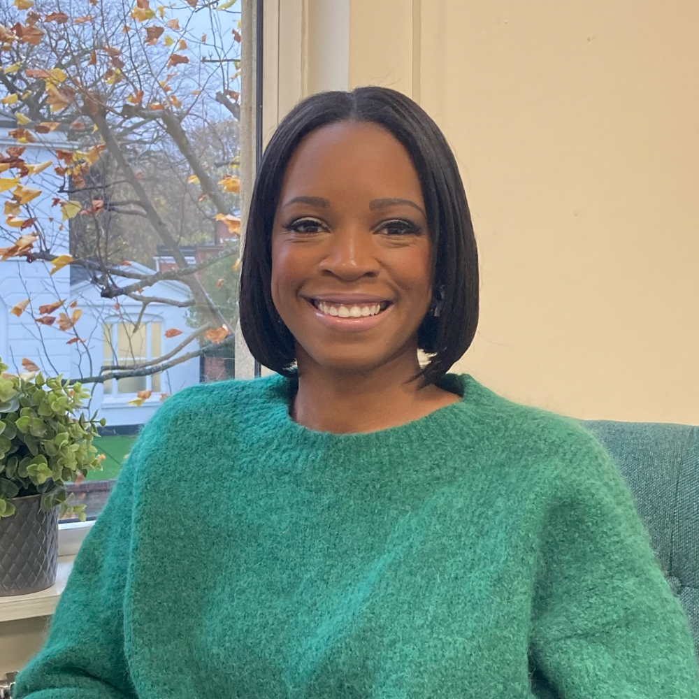 A woman with short black hair smiling, wearing a green sweater, sitting indoors near a window with a view of autumn trees and a house outside.