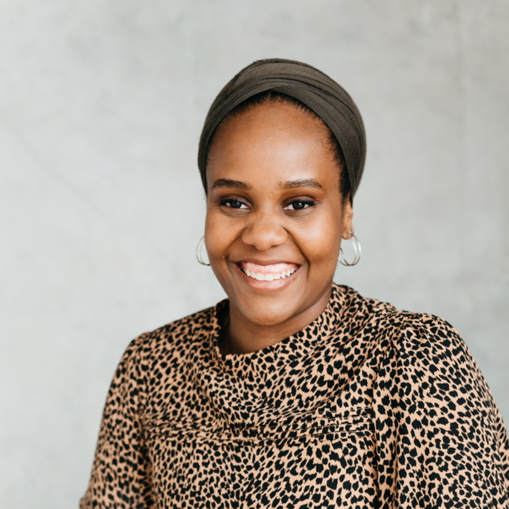 A smiling woman with dark skin, wearing a brown headwrap and leopard print top, against a plain neutral background.