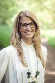 A smiling woman with long blonde hair wearing glasses and a white blouse, outdoors with trees in the background.