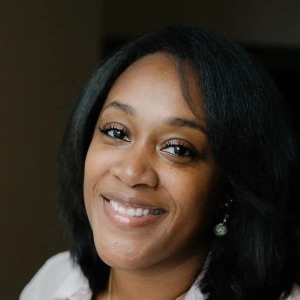 Close-up portrait of a smiling woman with shoulder-length black hair, wearing pearl earrings and a white top, against a dark background.